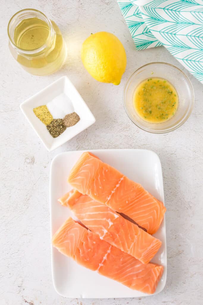 Overhead view of raw salmon fillets, seasonings, lemon, olive oil, and melted garlic butter laid out on a light surface.