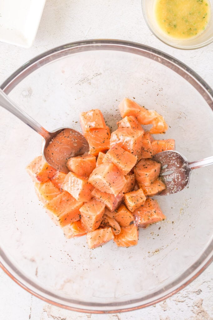 Cubes of raw salmon in a glass bowl being mixed with oil and seasonings using two metal spoons.