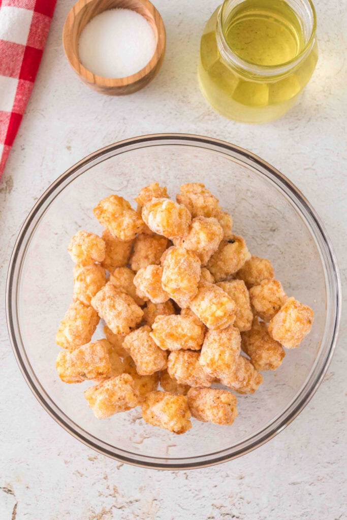 Frozen tater tots in a clear glass bowl with salt and oil sitting nearby on a light countertop.