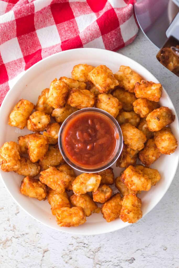 Overhead shot of a plate of tater tots with ketchup in the center and a red checkered cloth nearby.