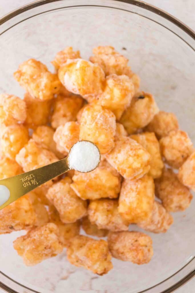 A measuring spoon of salt held over a bowl of frozen tater tots ready to be seasoned.