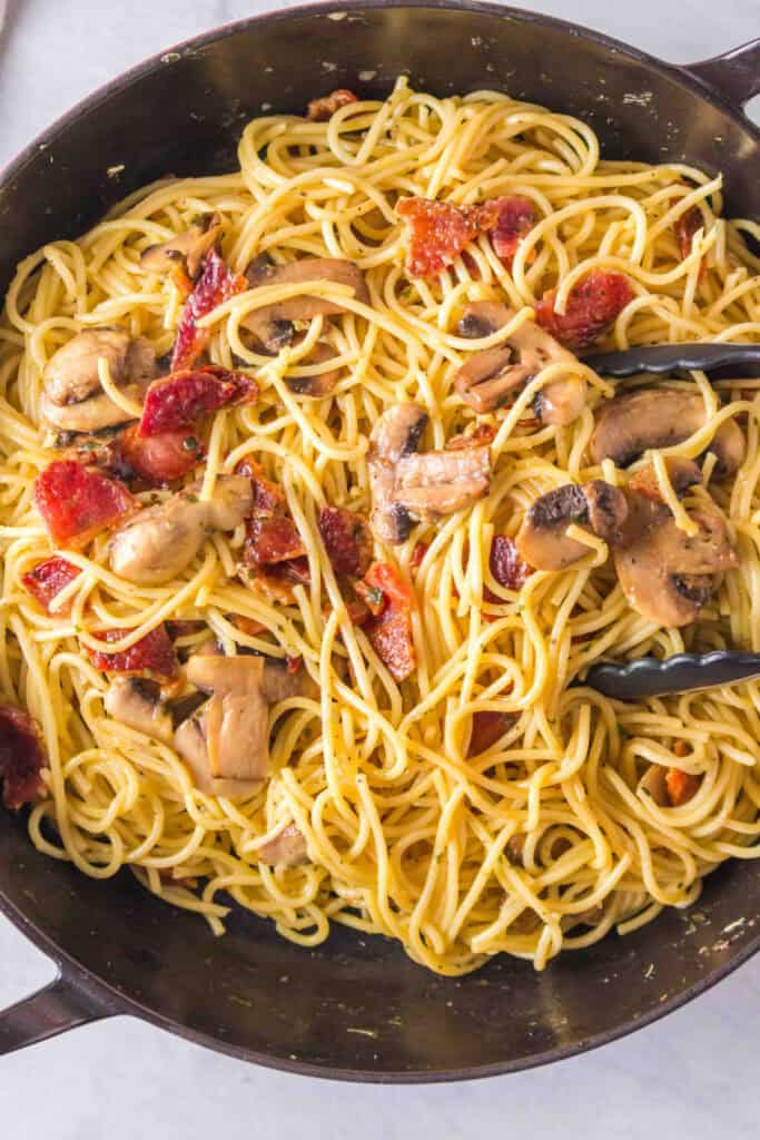 Spaghetti tossed with bacon and mushrooms in a skillet with tongs visible.