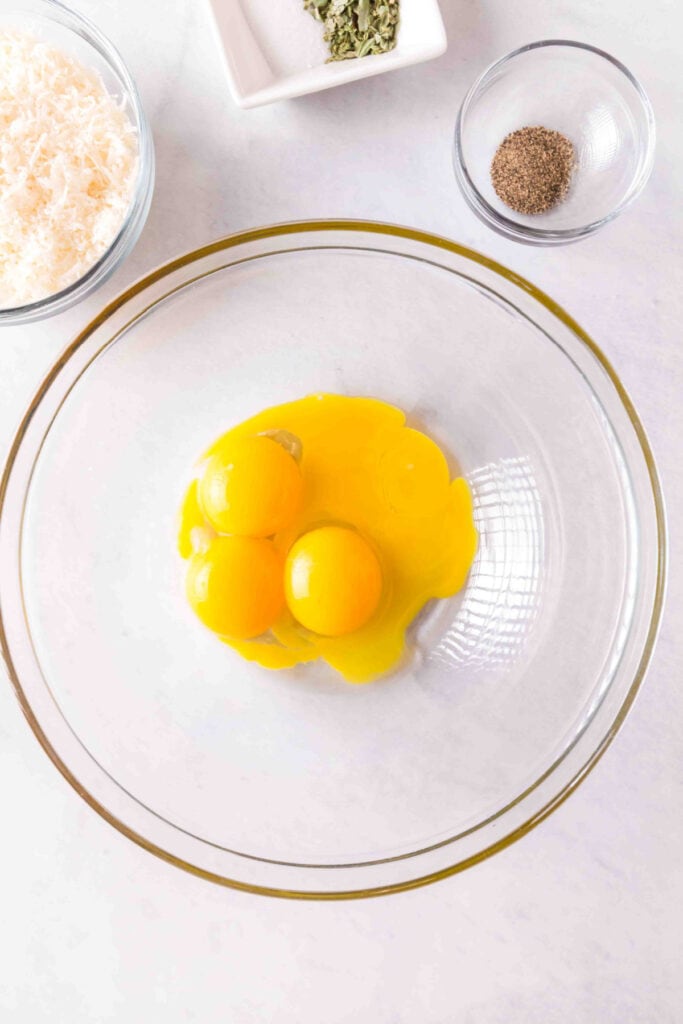Overhead view of three egg yolks in a clear glass bowl on a white surface with grated cheese, basil, and pepper nearby.