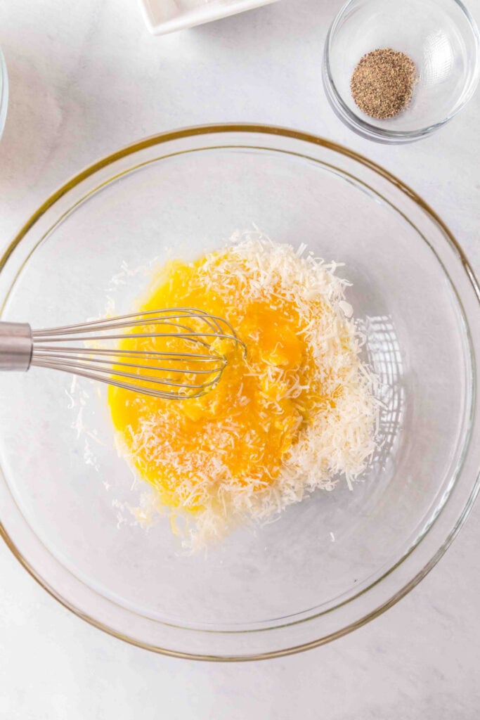 Egg yolks in a glass bowl topped with freshly grated cheese and a whisk resting inside.