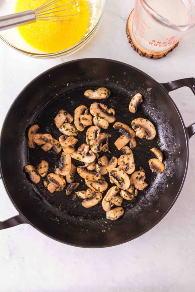 Sliced mushrooms sautéing in a black skillet with herbs visible on the surface.