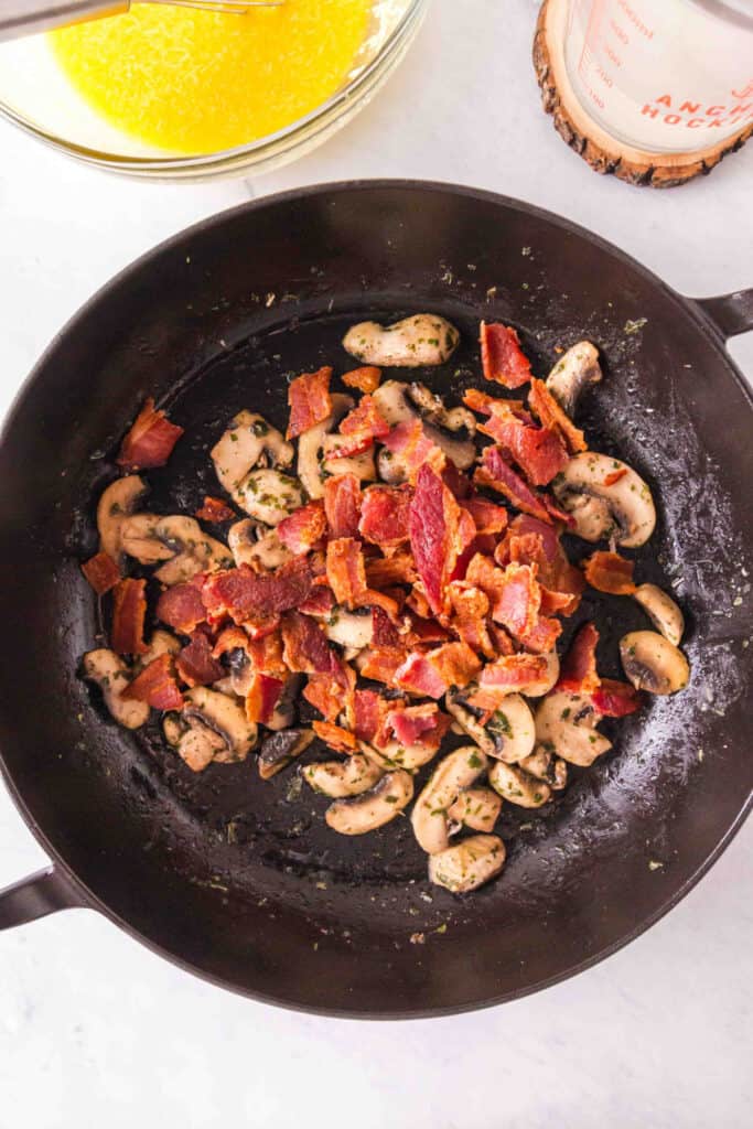 Cooked sliced mushrooms in a skillet topped with crumbled cooked bacon pieces.