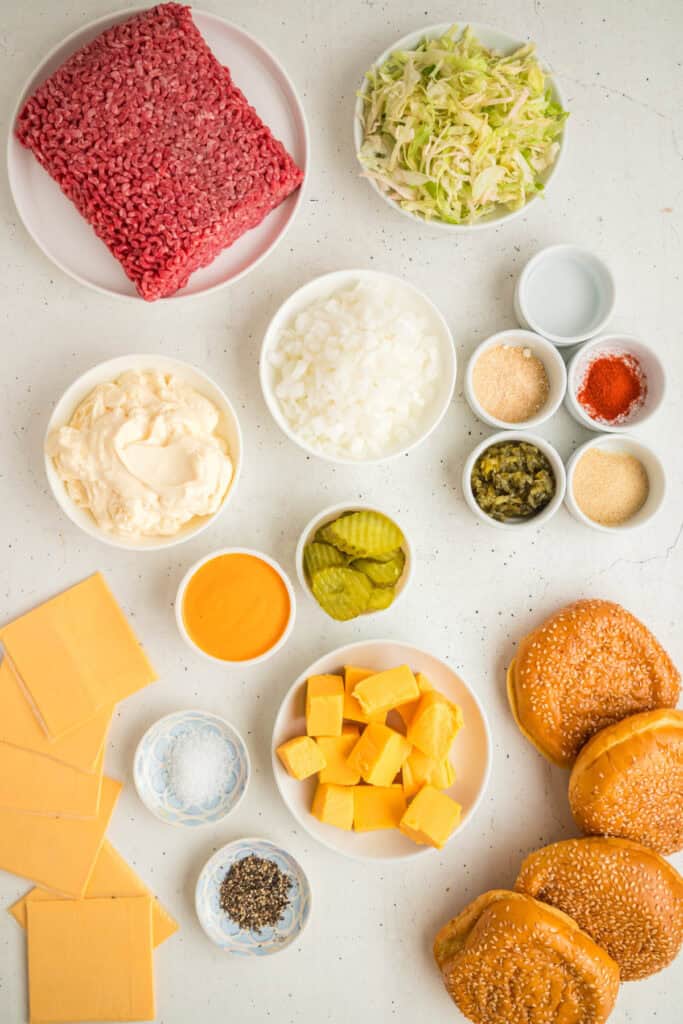 Overhead view of all Big Mac Sloppy Joes ingredients, including ground beef, lettuce, onions, cheese, pickles, mayonnaise, and sesame seed buns arranged on a white surface.