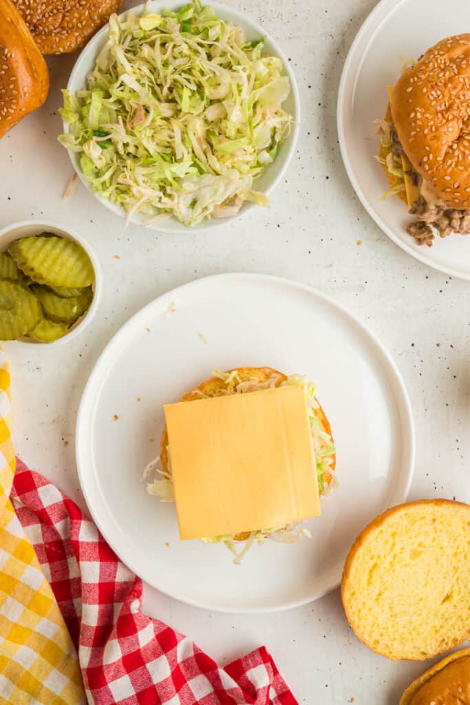 Assembling Big Mac Sloppy Joes with shredded lettuce and a slice of American cheese on a bun, surrounded by pickles and extra buns.