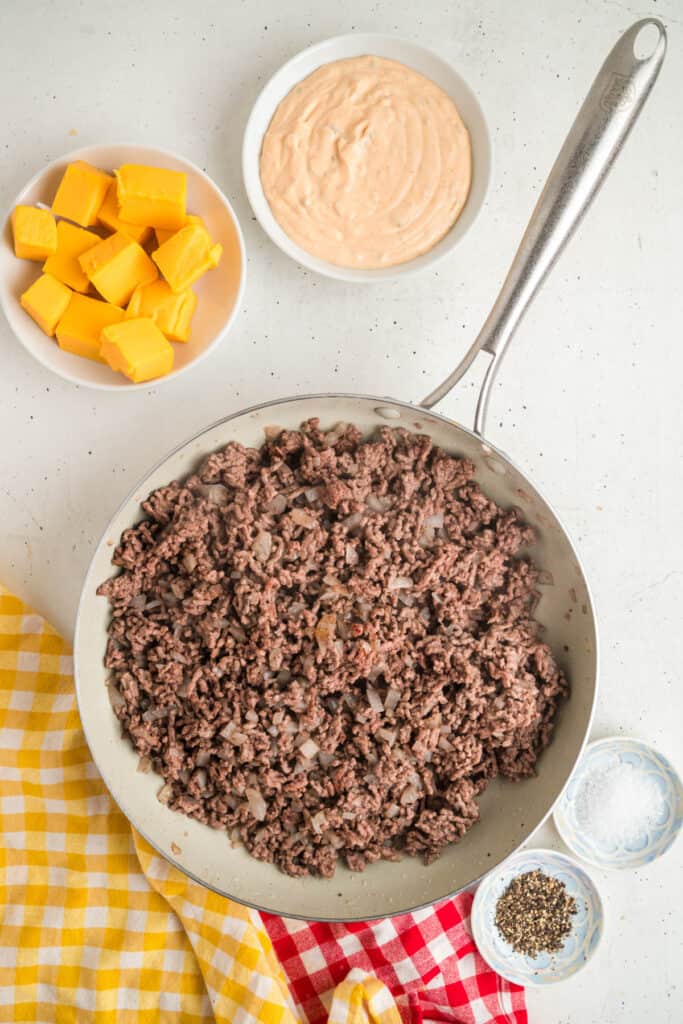 Cooked ground beef and onion mixture in a skillet with bowls of cheese cubes and Big Mac sauce nearby.