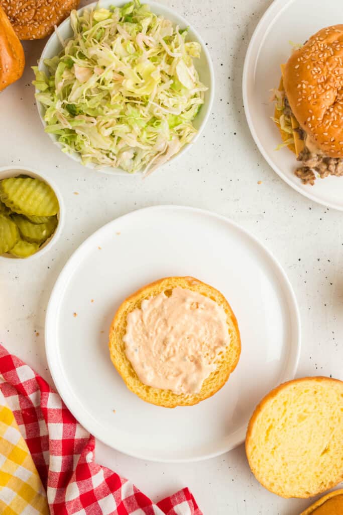 Bottom hamburger bun spread with Big Mac sauce next to shredded lettuce and pickles for assembling Big Mac Sloppy Joes.