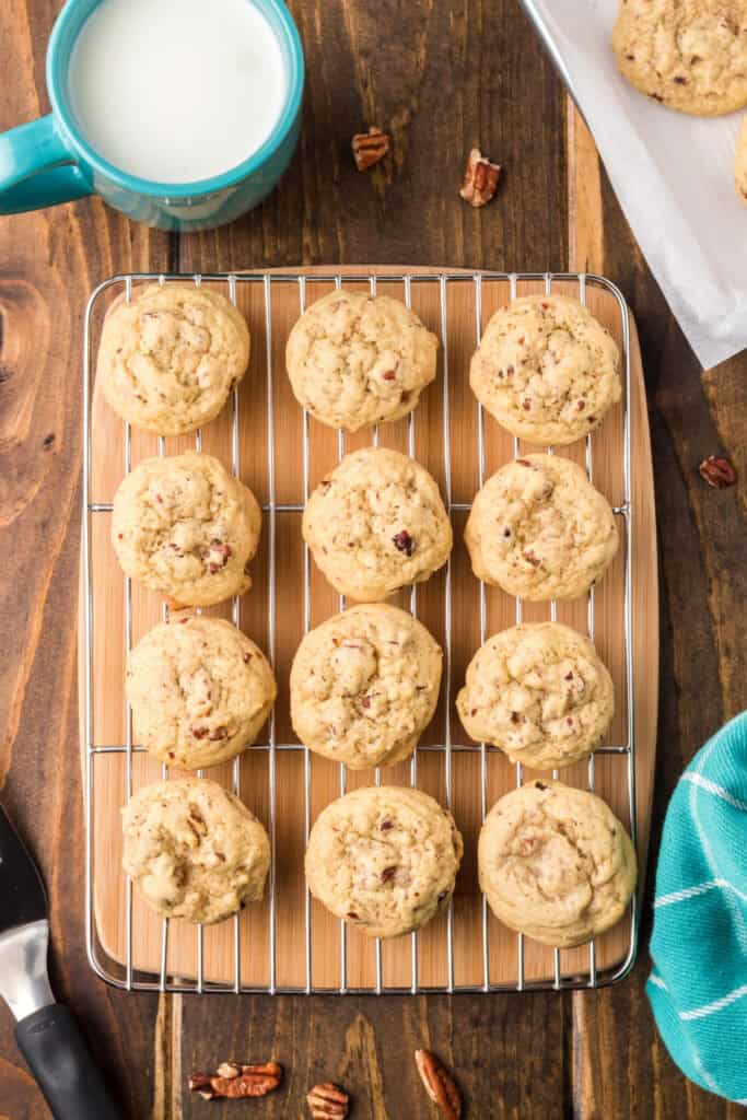 Butter Pecan Cookies cooling on a wire rack beside a turquoise mug of milk and scattered pecans on a wooden table.