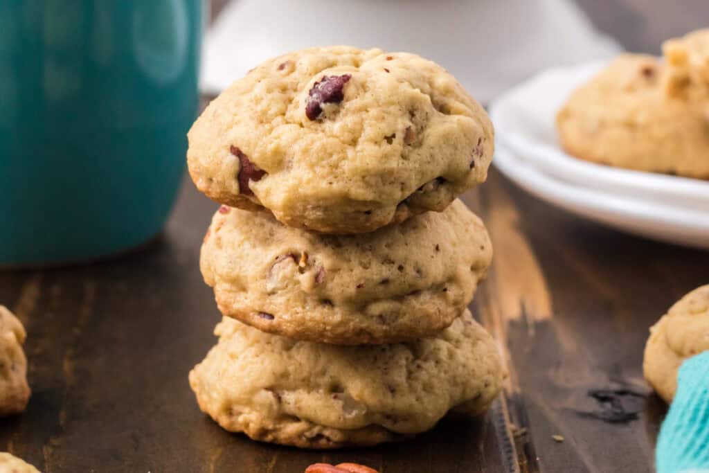 Stack of three Butter Pecan Cookies showing soft centers and chopped pecans on a wooden table beside a teal mug.