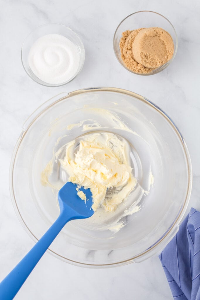 Butter creamed in a glass mixing bowl with a blue spatula beside bowls of brown and granulated sugar for Butter Pecan Cookies.