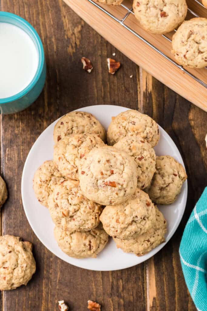 Plate of freshly baked Butter Pecan Cookies beside a turquoise mug of milk and a cooling rack on a wooden surface.