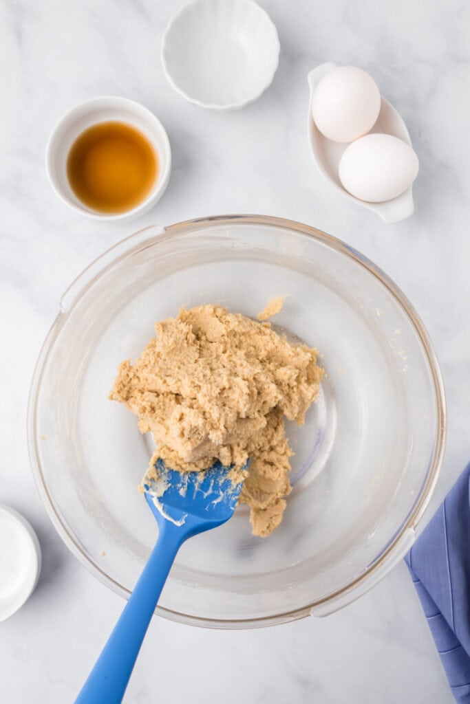 Cookie dough mixed with butter and sugars in a clear bowl with eggs and vanilla extract in the background for Butter Pecan Cookies.