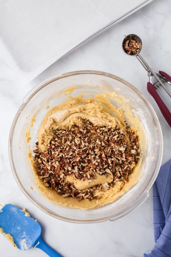 Chopped pecans added into the cookie dough mixture in a glass bowl with a blue spatula for Butter Pecan Cookies.