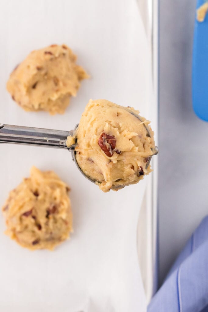 Cookie scoop holding a portion of Butter Pecan Cookies dough with pecan pieces visible over a lined baking sheet.