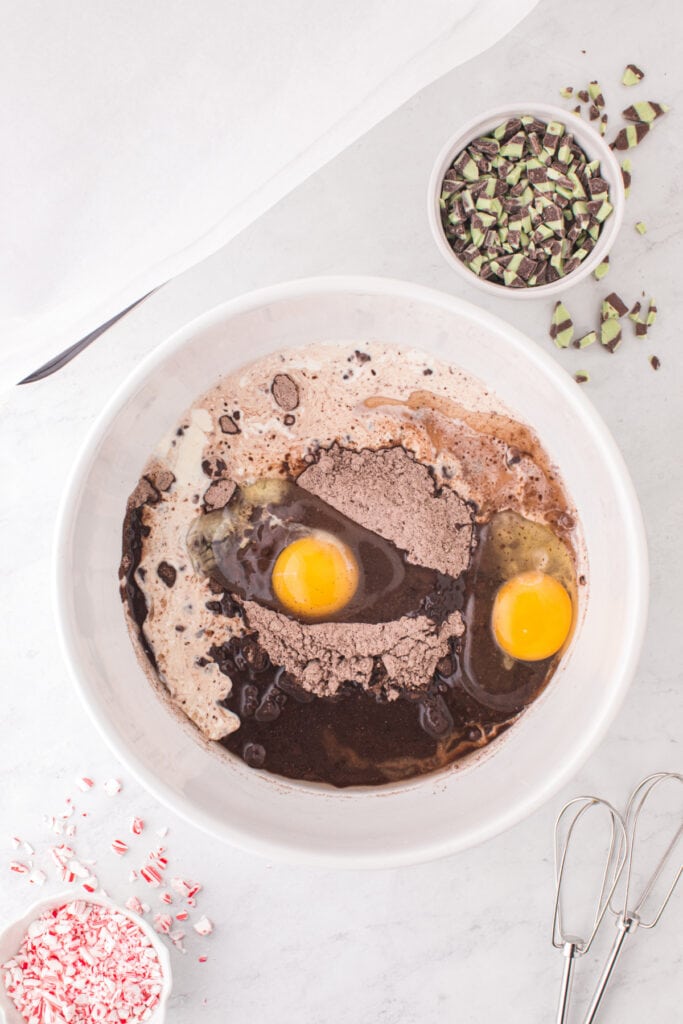 A white mixing bowl filled with brownie mix, eggs, oil and cream before mixing, surrounded by crushed candy canes and mint baking chips.