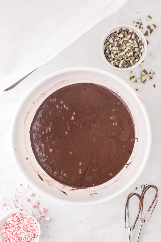 A bowl of freshly mixed chocolate brownie batter with smooth texture, with bowls of mint baking chips and crushed candy canes nearby.