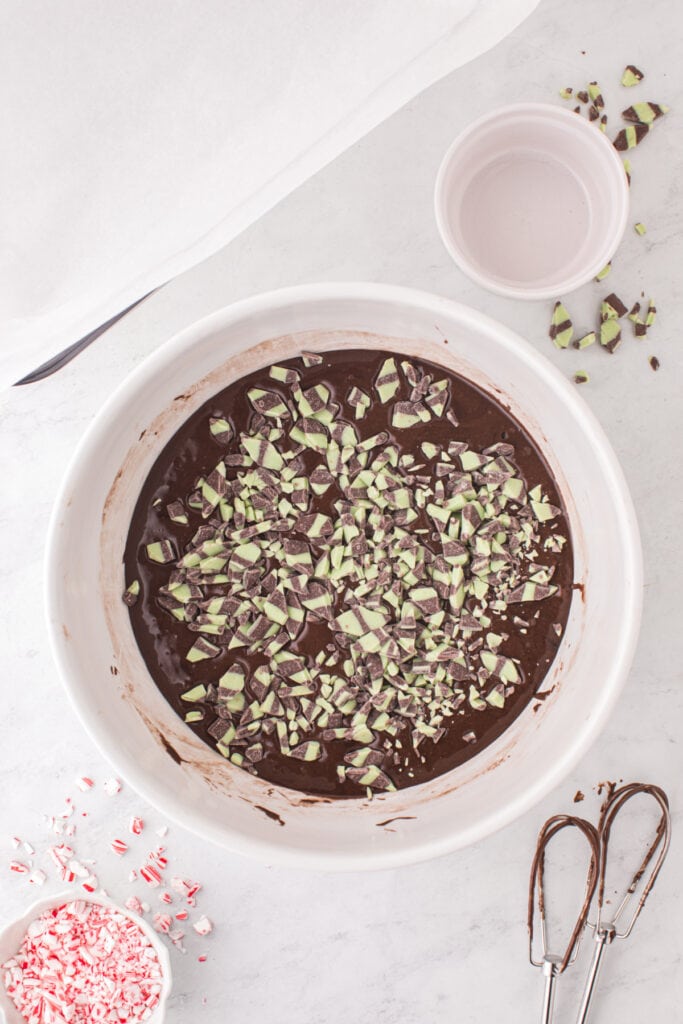Mint fudge baking chips scattered on top of unblended brownie batter in a large white mixing bowl.