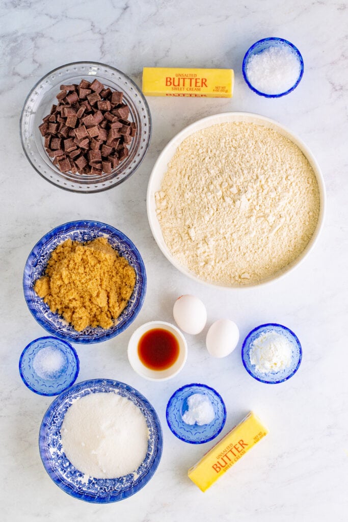 Overhead view of cookie ingredients arranged in bowls, including flour, sugars, butter, eggs, vanilla, and chocolate chunks.