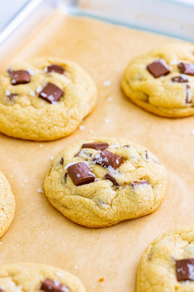 Close up of warm cookies with chocolate chunks and flaky sea salt cooling on a parchment lined baking sheet.