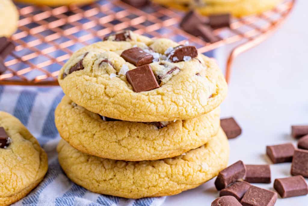 Stack of three soft cookies with chocolate chunks on top, sitting on a cloth with extra chocolate pieces nearby.