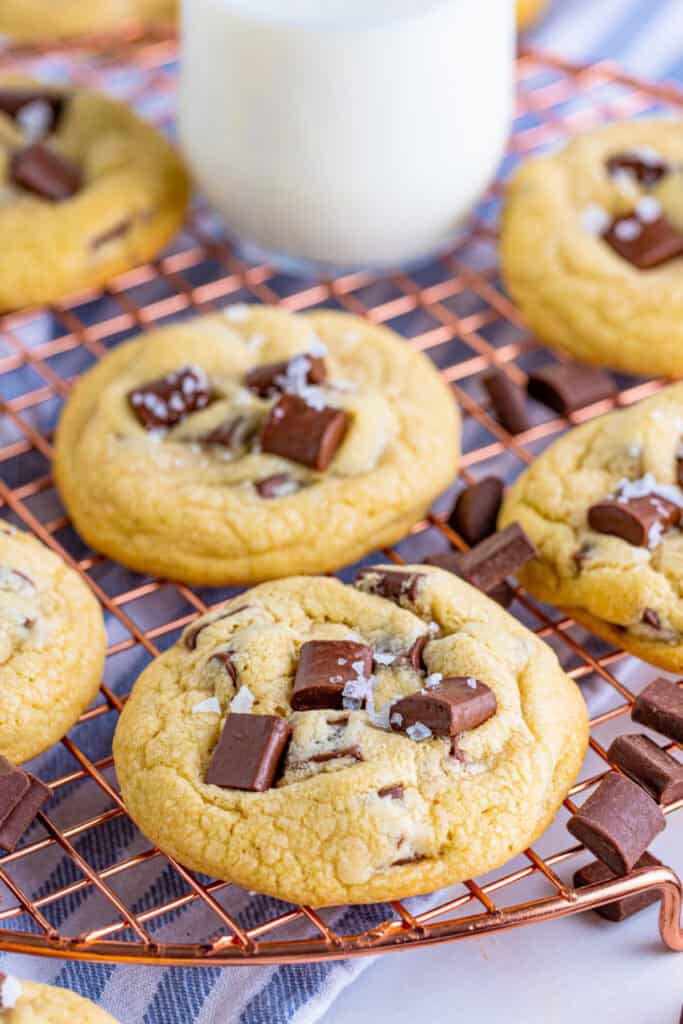 Cookies topped with chocolate chunks and flaky salt cooling on a copper rack with a glass of milk in the background.
