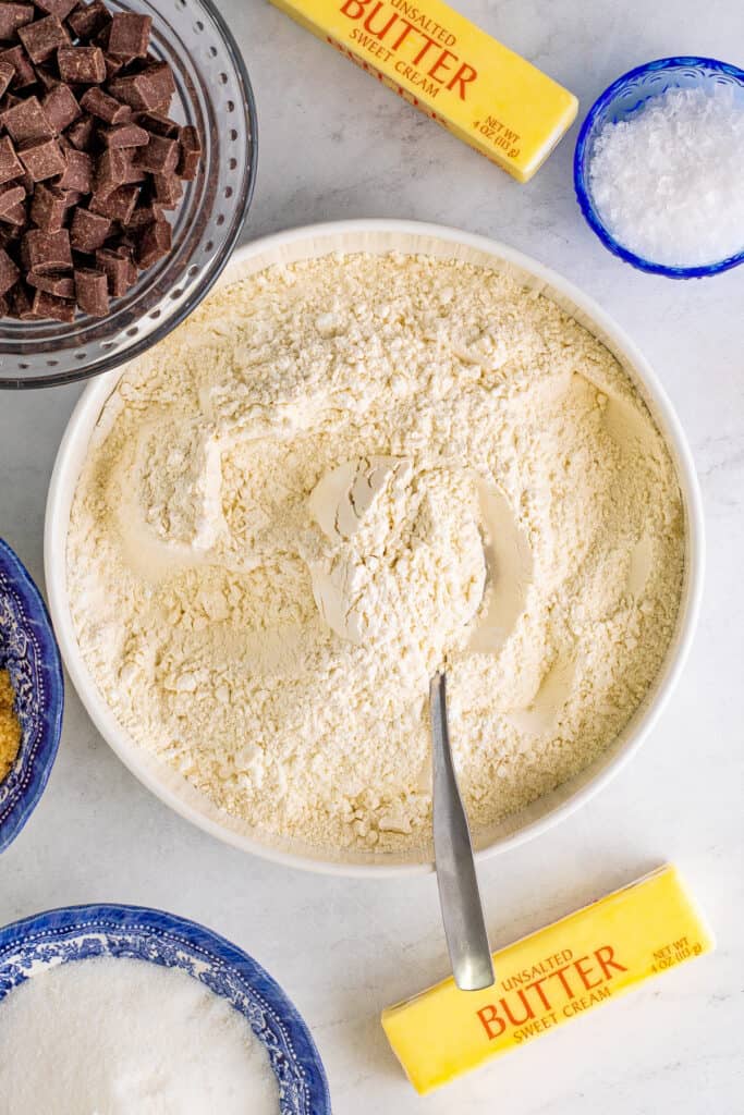 Bowl of flour mixture with a spoon resting in it, surrounded by chocolate chunks, butter, and other ingredients.