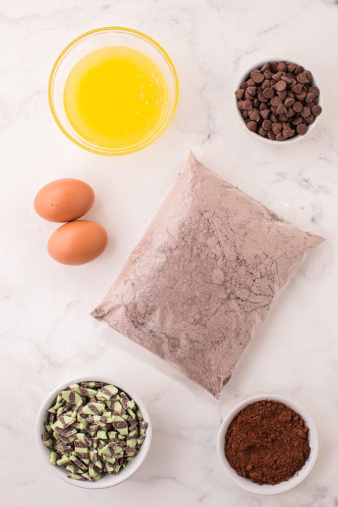 Ingredients for Chocolate Mint Chip Cookies including eggs, melted butter, cake mix, cocoa powder, milk chocolate chips, and mint chips arranged on a white marble surface.