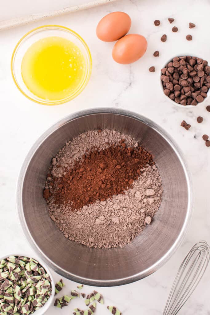Mixing bowl with chocolate cake mix and cocoa powder surrounded by baking ingredients like eggs, melted butter, and mint chips.