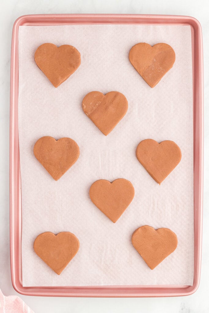 Heart shaped unbaked chocolate cookies arranged evenly on a parchment lined baking sheet.