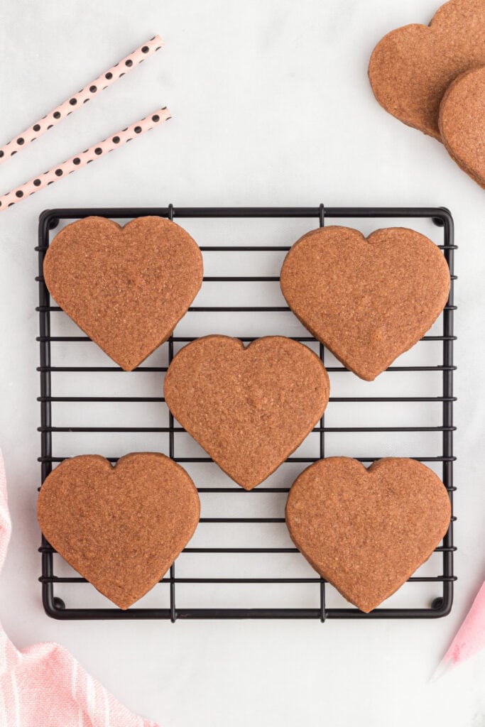 Cooled heart shaped chocolate cookies on a black wire rack with pink straws and additional cookies in the background.