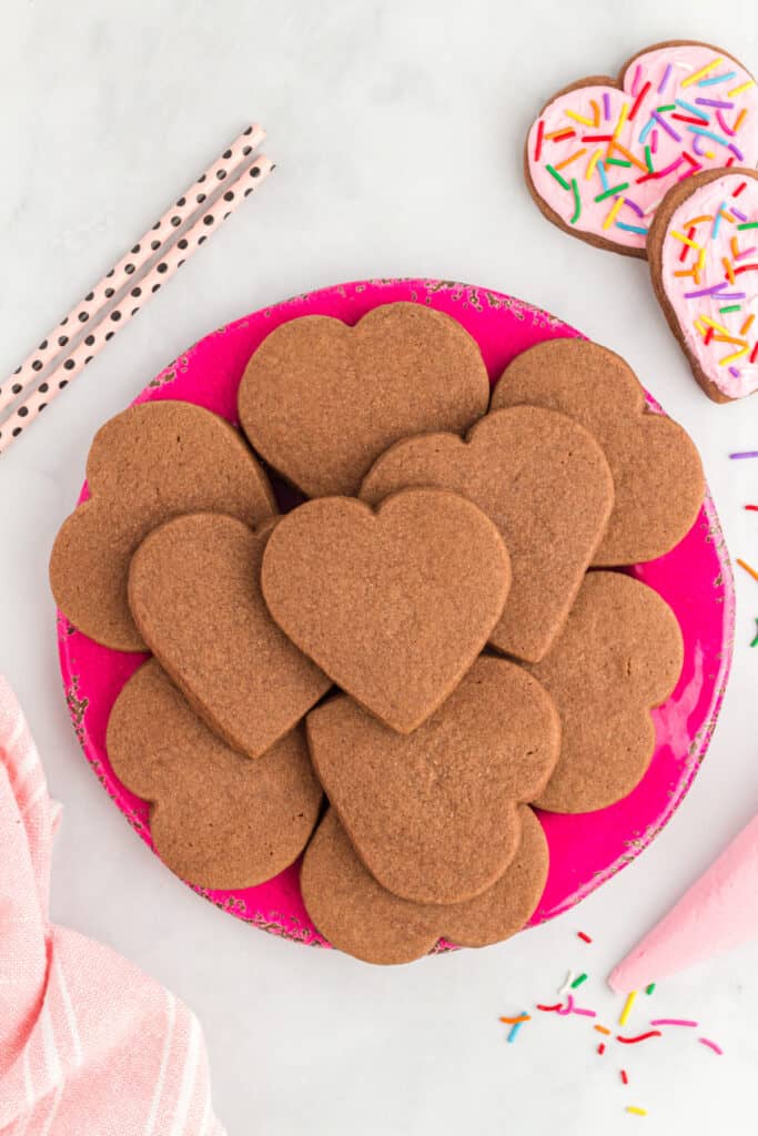 Overhead view of unfrosted heart shaped chocolate cookies arranged on a bright pink plate with sprinkles and straws nearby.