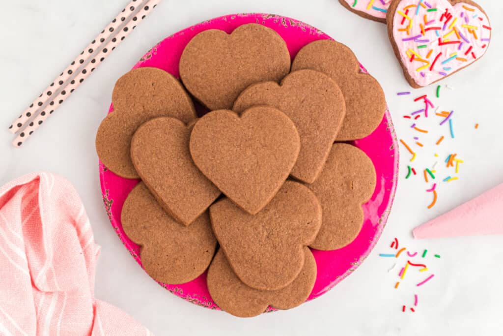 Wide overhead shot of heart shaped chocolate cookies on a pink plate surrounded by sprinkles, straws, and a piping bag.