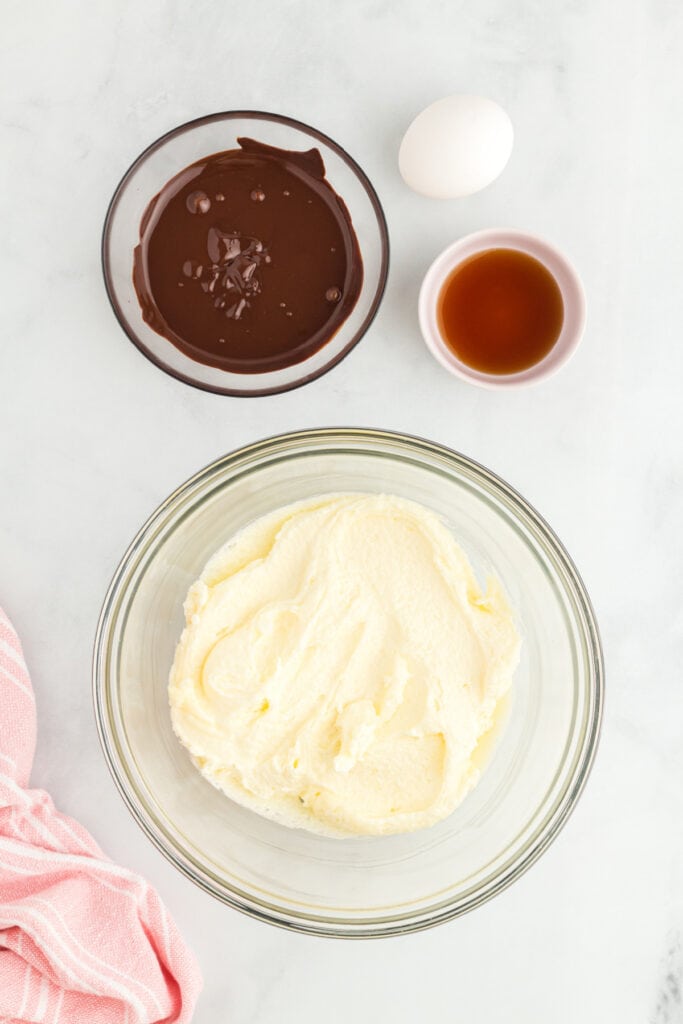 Creamed butter and sugar mixture in a glass bowl with melted chocolate, egg, and vanilla nearby on a white surface.