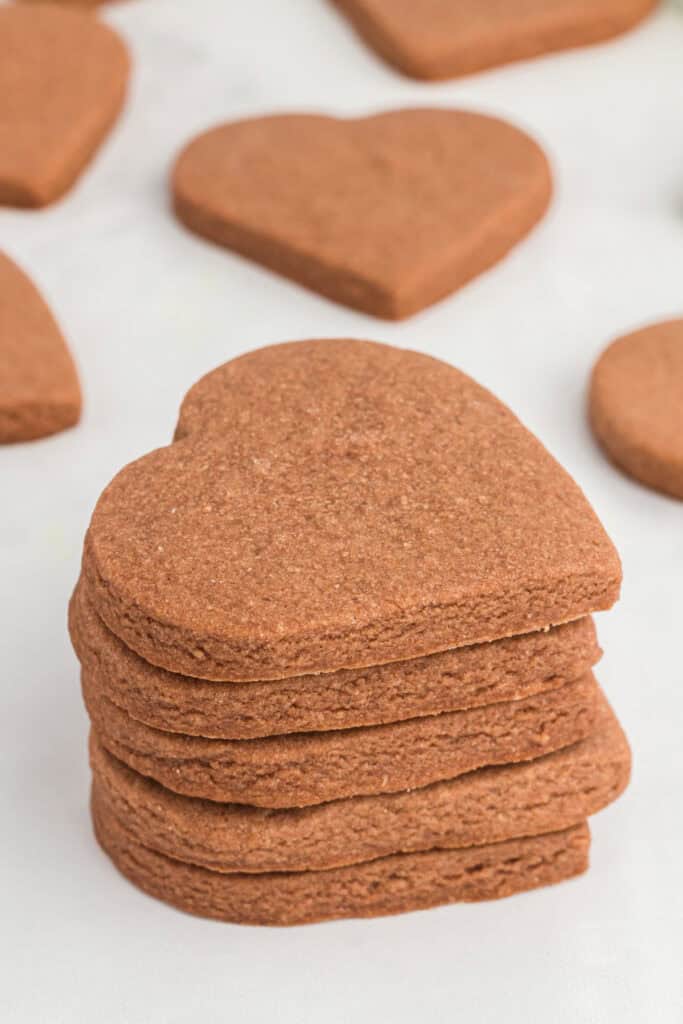 Stack of heart shaped chocolate cookies with more cookies scattered in the background on a white surface.