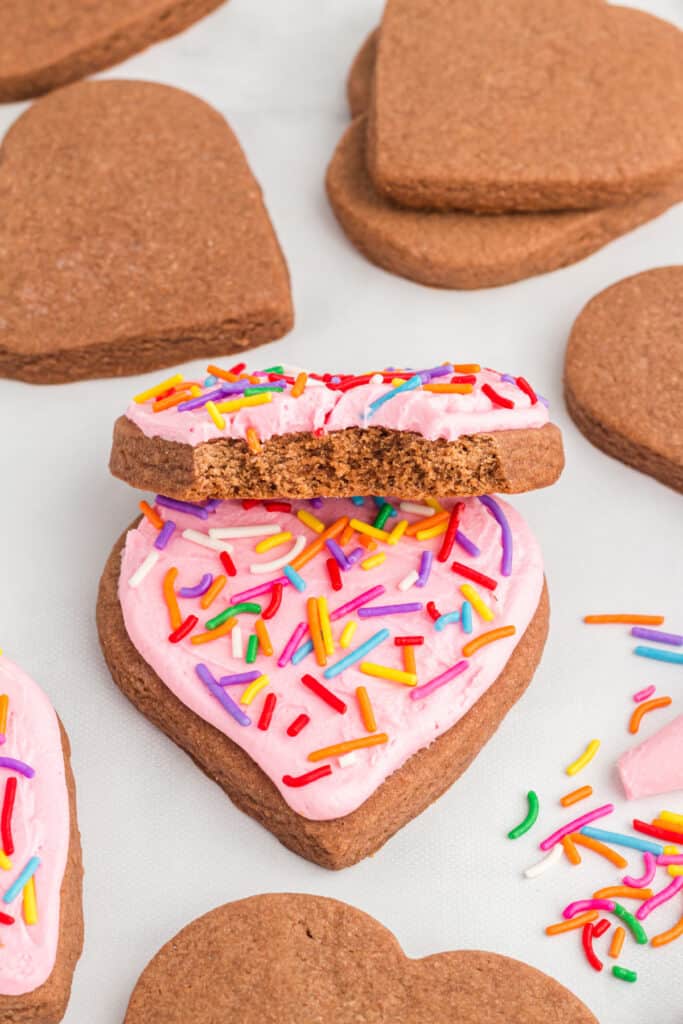 Frosted heart shaped chocolate cookie with pink frosting and rainbow sprinkles, with a bite taken out of the top cookie.