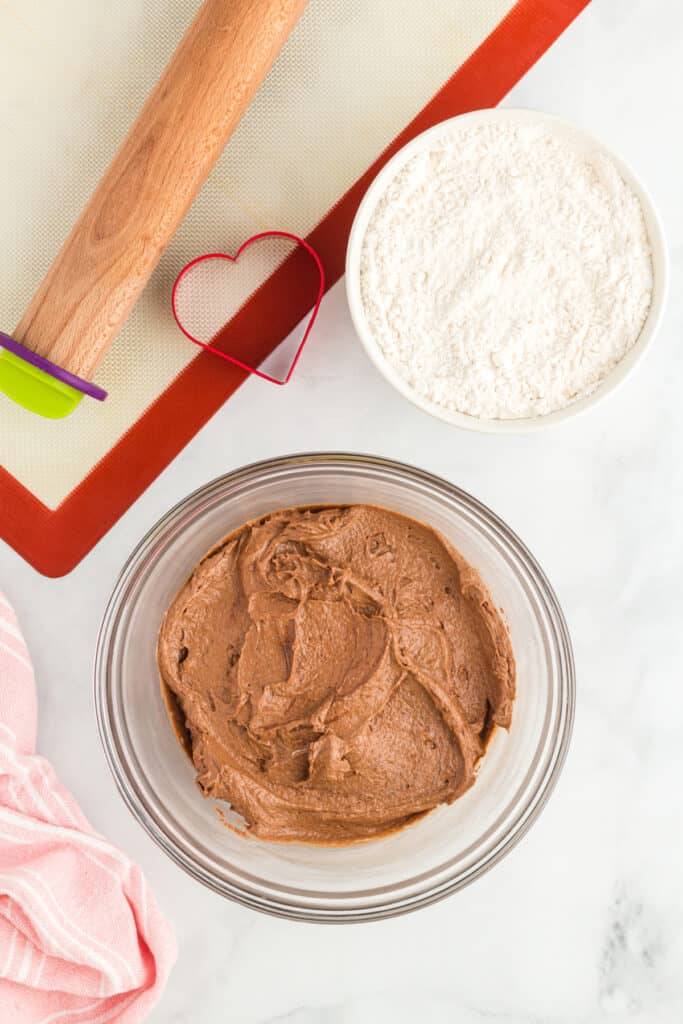 Chocolate cookie dough fully mixed in a glass bowl with a rolling pin, silicone mat, and heart cookie cutter nearby.