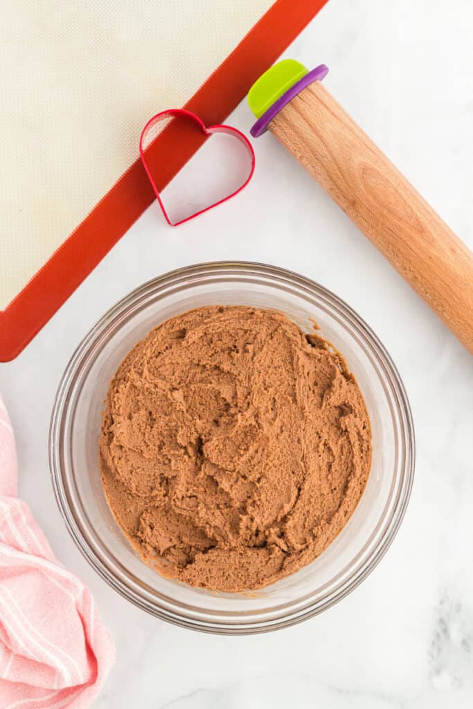 Thick chocolate cookie dough in a bowl next to a rolling pin and heart cookie cutter on a silicone baking mat.