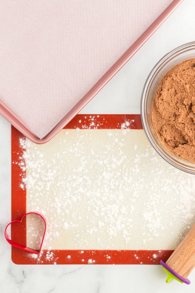 Silicone mat lightly coated with flour next to a bowl of chocolate cookie dough and a parchment lined baking sheet.