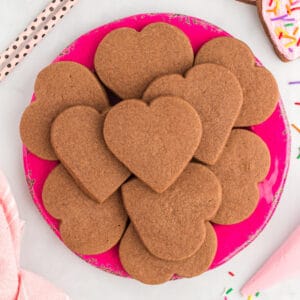 Square overhead view of heart shaped chocolate cookies arranged neatly on a bright pink plate with sprinkles nearby.