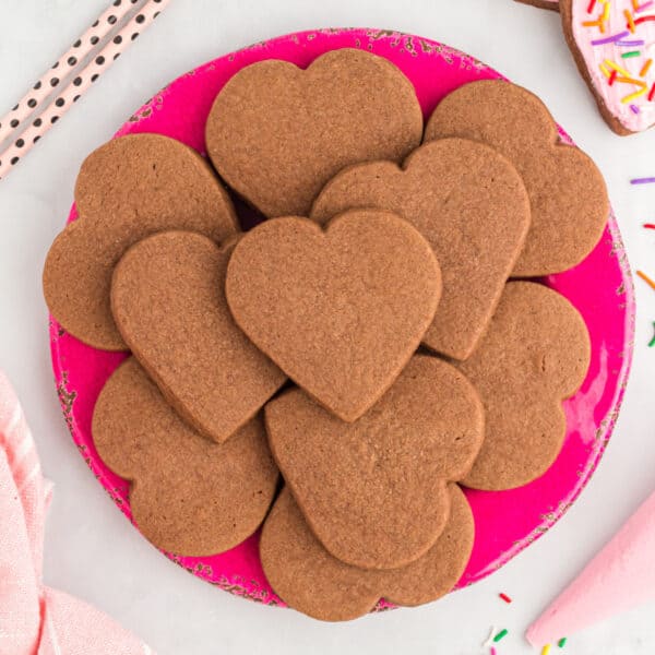 Square overhead view of heart shaped chocolate cookies arranged neatly on a bright pink plate with sprinkles nearby.