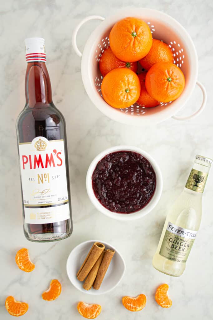Ingredients for Cranberry Pimm’s Cocktail including Pimm’s No.1 bottle, clementines, cranberry sauce, ginger beer, and cinnamon sticks on a marble surface.