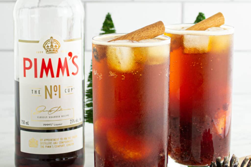 Two Cranberry Pimm’s Cocktails with ice and cinnamon sticks beside a Pimm’s No.1 bottle, creating a cozy holiday scene.