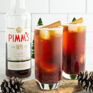 Two glasses of Cranberry Pimm’s Cocktail garnished with cinnamon sticks, surrounded by pinecones and festive décor.