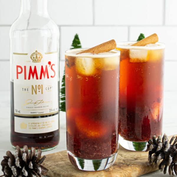 Two glasses of Cranberry Pimm’s Cocktail garnished with cinnamon sticks, surrounded by pinecones and festive décor.