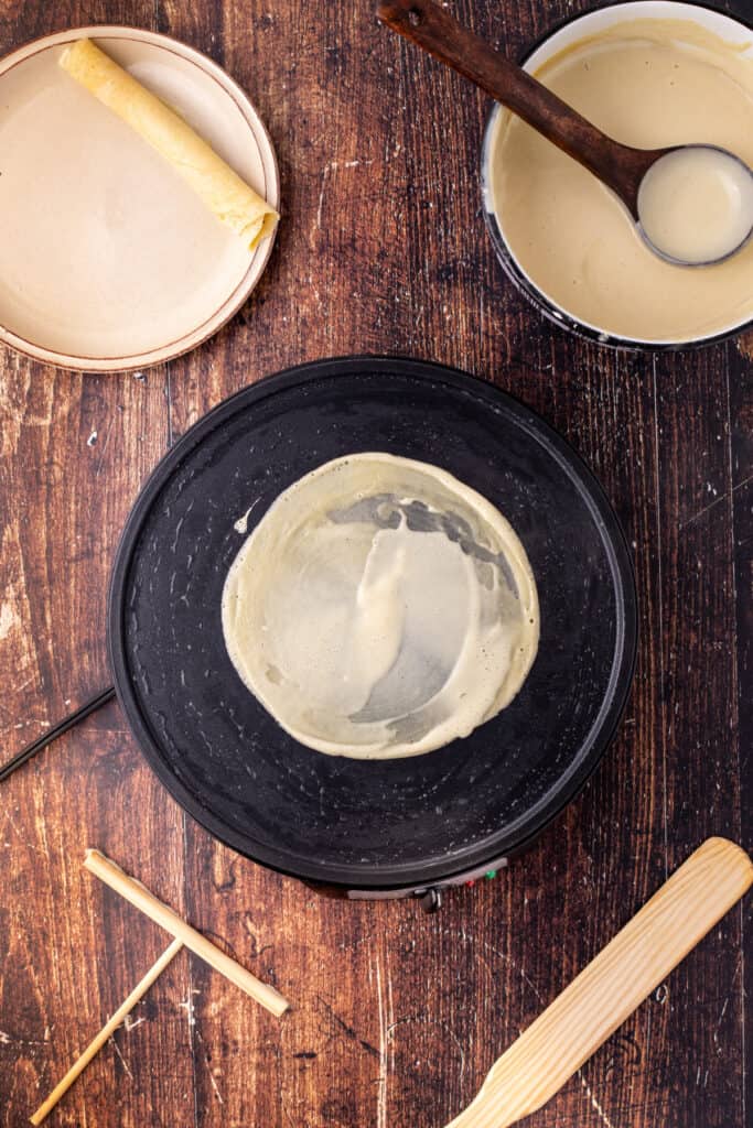 Finished eggnog crepe cooking on the pan with one rolled crepe on a plate in the background, ready for serving.