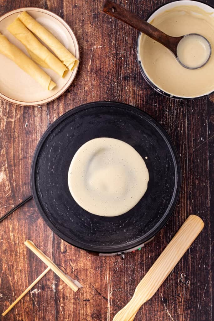 A crêpe cooking on a hot plate with a ladle of eggnog batter nearby and rolled crêpes on a plate beside it.