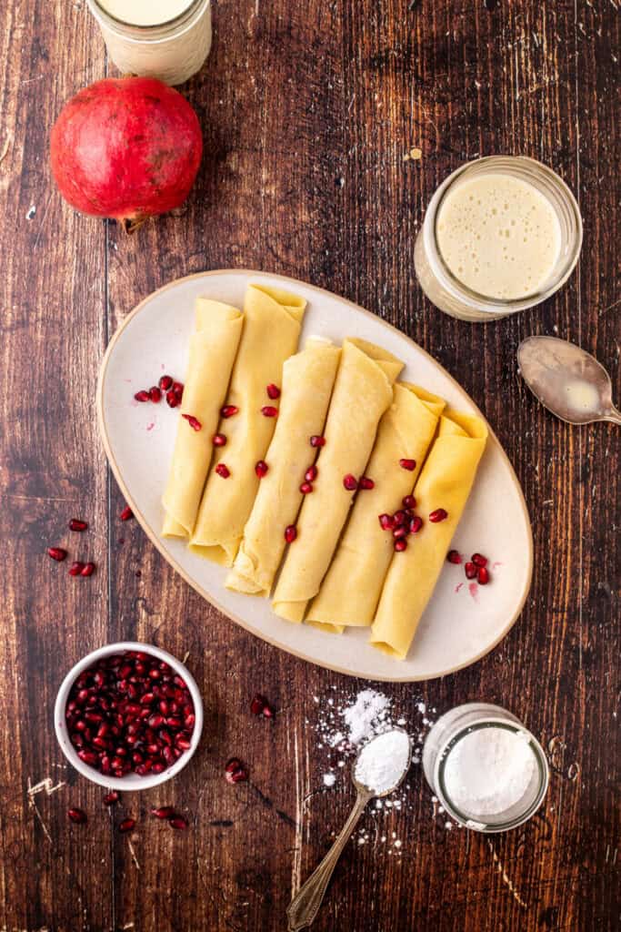 A plate of plain rolled eggnog crêpes on a rustic wood table, surrounded by pomegranate seeds, a jar of eggnog sauce, and a spoon with powdered sugar.